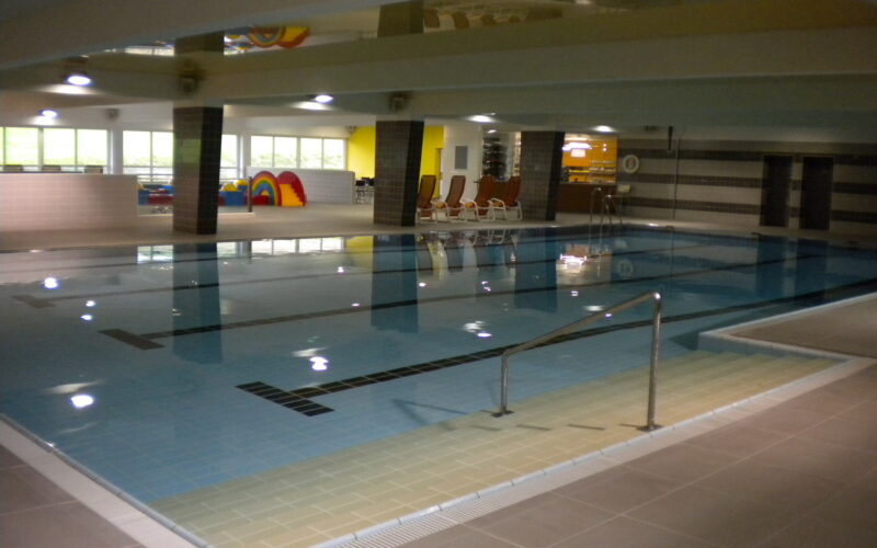 Innenpool mit klarem Wasser, Poolleiter, gefliestem Boden und Liegestühlen im Hintergrund unter fluoreszierender Beleuchtung.