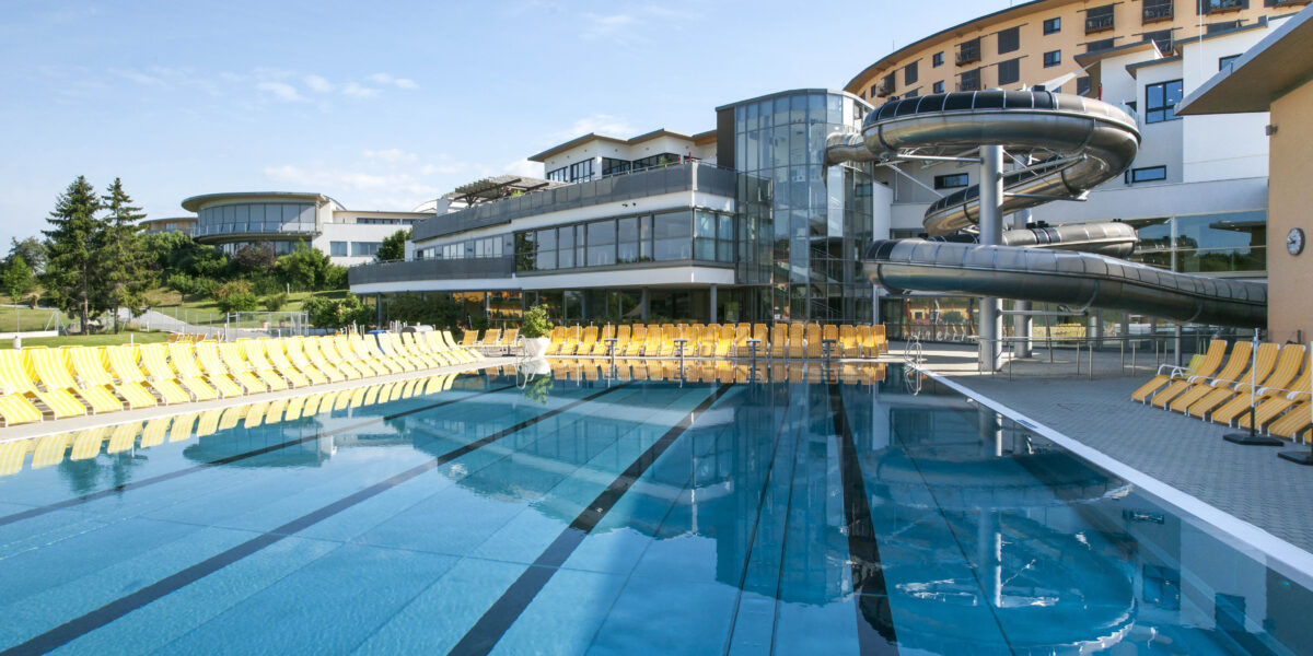 Freibad mit markierten Bahnen, umgeben von gelben Liegestühlen, neben einem modernen Gebäude mit großen Wasserrutschen unter strahlend blauem Himmel.