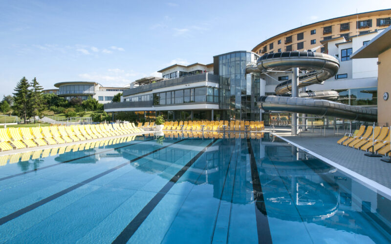 Freibad mit markierten Bahnen, umgeben von gelben Liegestühlen, neben einem modernen Gebäude mit großen Wasserrutschen unter strahlend blauem Himmel.