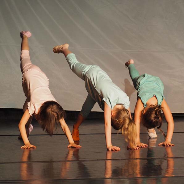 Drei kleine Kinder führen auf der Bühne eine Tanzeinlage auf, wobei jedes auf einem Bein und einer Hand balanciert und das andere Bein nach hinten angehoben ist.