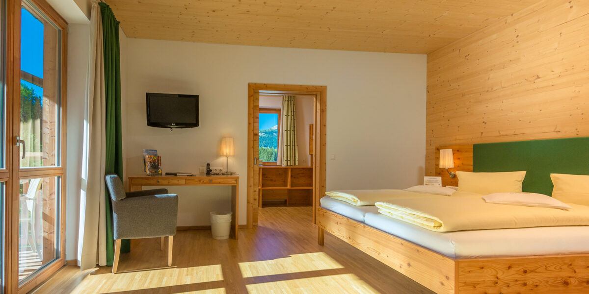 Ein helles Hotelzimmer mit einem Doppelbett, Holzmöbeln, einem Schreibtisch mit Stuhl, einem an der Wand montierten Fernseher und einer Tür, die zum Badezimmer führt. Durch die großen Fenster strömt Sonnenlicht herein.