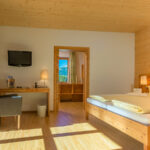 Ein helles Hotelzimmer mit einem Doppelbett, Holzmöbeln, einem Schreibtisch mit Stuhl, einem an der Wand montierten Fernseher und einer Tür, die zum Badezimmer führt. Durch die großen Fenster strömt Sonnenlicht herein.