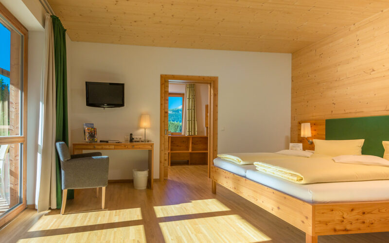 Ein helles Hotelzimmer mit einem Doppelbett, Holzmöbeln, einem Schreibtisch mit Stuhl, einem an der Wand montierten Fernseher und einer Tür, die zum Badezimmer führt. Durch die großen Fenster strömt Sonnenlicht herein.