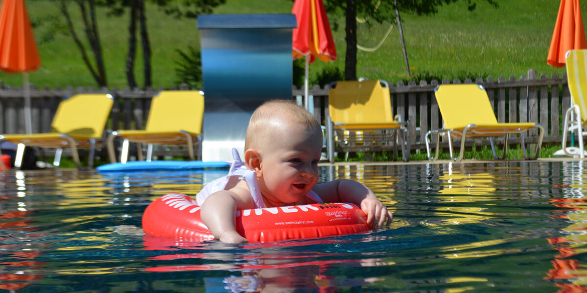 Ein Baby sitzt in einem roten aufblasbaren Ring, der in einem Pool schwimmt, umgeben von gelben Liegestühlen und orangefarbenen Sonnenschirmen an einem sonnigen Tag.
