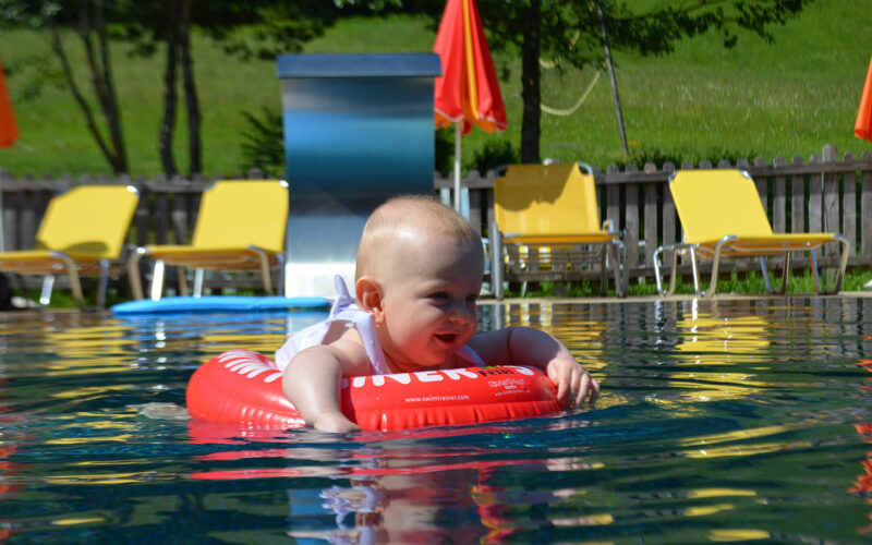 Ein Baby sitzt in einem roten aufblasbaren Ring, der in einem Pool schwimmt, umgeben von gelben Liegestühlen und orangefarbenen Sonnenschirmen an einem sonnigen Tag.