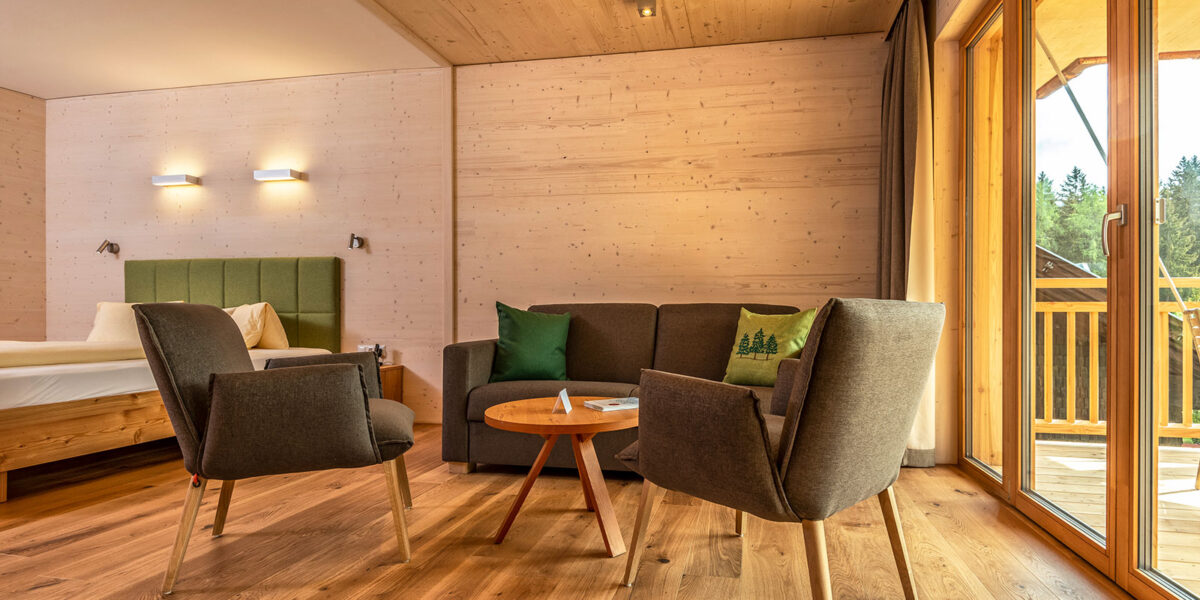 Ein modernes Hotelzimmer mit Holzfußboden, einem Bett, einem grünen Sofa, zwei grauen Sesseln um einen runden Tisch und großen Fenstern, die auf einen Balkon mit Blick auf den Wald führen.
