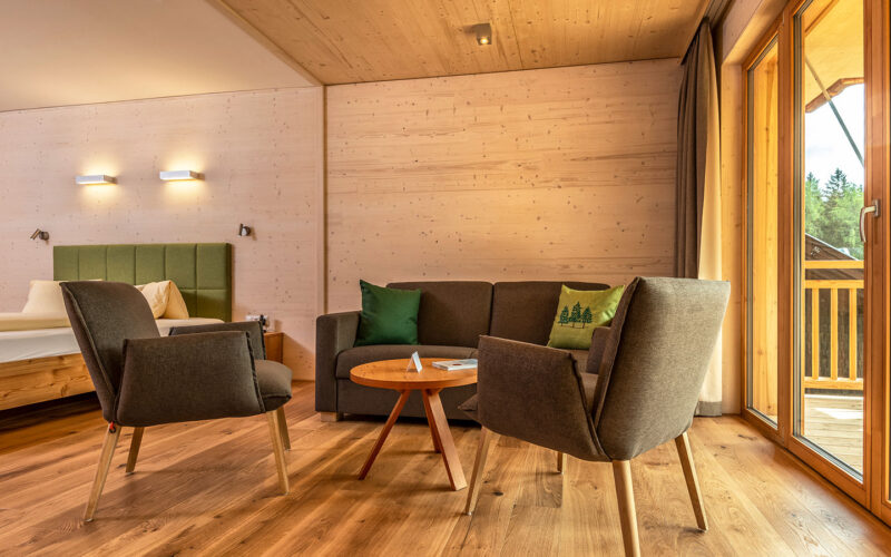 Ein modernes Hotelzimmer mit Holzfußboden, einem Bett, einem grünen Sofa, zwei grauen Sesseln um einen runden Tisch und großen Fenstern, die auf einen Balkon mit Blick auf den Wald führen.