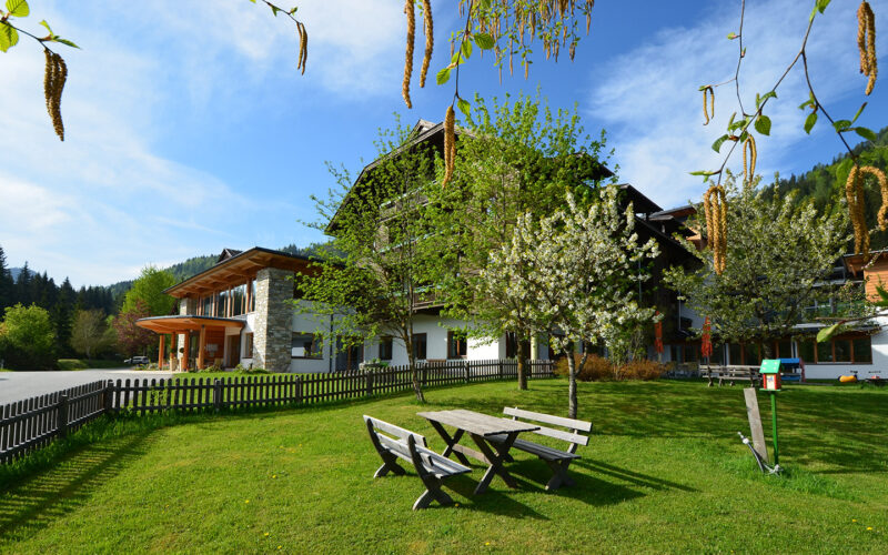 Hinter einer eingezäunten Wiese mit Picknicktischen und blühenden Bäumen steht unter blauem Himmel ein Holzgebäude mit Schrägdach.