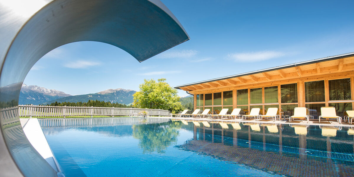 Außenschwimmbad mit Wasserrutsche, Sonnenliegen und einem modernen Holzgebäude vor der Kulisse der Berge und des blauen Himmels.
