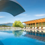 Außenschwimmbad mit Wasserrutsche, Sonnenliegen und einem modernen Holzgebäude vor der Kulisse der Berge und des blauen Himmels.