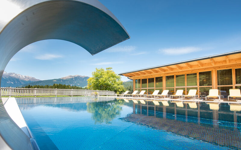 Außenschwimmbad mit Wasserrutsche, Sonnenliegen und einem modernen Holzgebäude vor der Kulisse der Berge und des blauen Himmels.