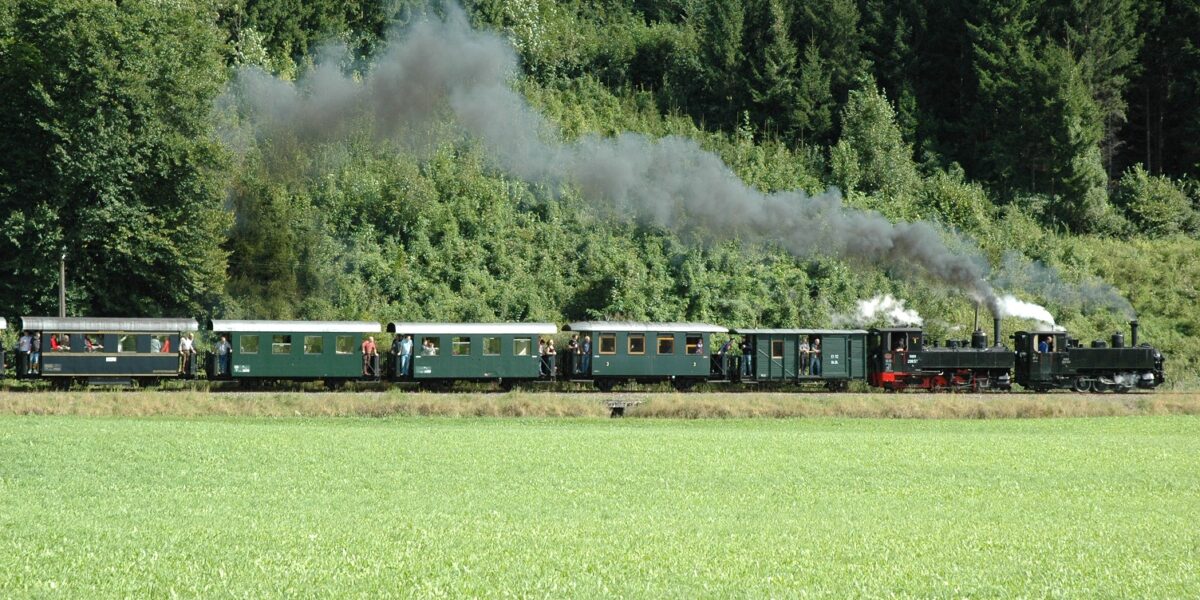 Ein alter Dampfzug mit grünen Waggons fährt durch eine Grasfläche mit Bäumen und Wald im Hintergrund und stößt schwarzen Rauch aus.