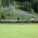 Ein alter Dampfzug mit grünen Waggons fährt durch eine Grasfläche mit Bäumen und Wald im Hintergrund und stößt schwarzen Rauch aus.