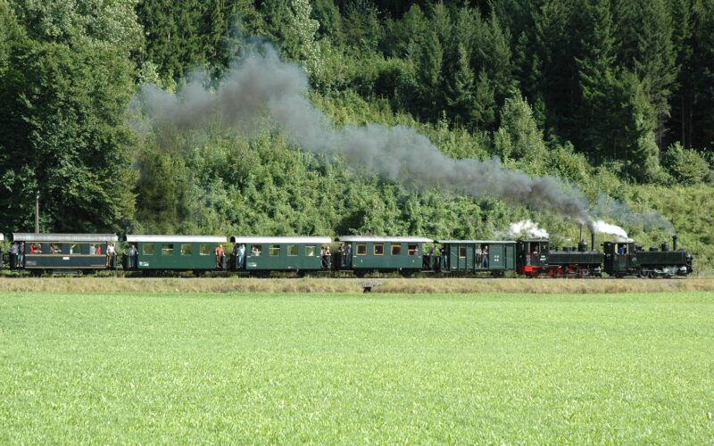 Ein alter Dampfzug mit grünen Waggons fährt durch eine Grasfläche mit Bäumen und Wald im Hintergrund und stößt schwarzen Rauch aus.