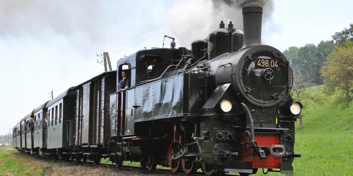 Eine schwarze Dampflok mit der Nummer 498.04 zieht Personenwaggons über ein Gleisnetz und stößt in einer ländlichen Gegend Rauch aus ihrem Schornstein.