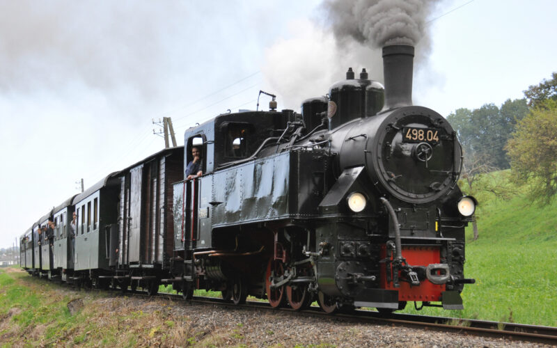 Eine schwarze Dampflok mit der Nummer 498.04 zieht Personenwaggons über ein Gleisnetz und stößt in einer ländlichen Gegend Rauch aus ihrem Schornstein.