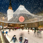 Schlittschuhlaufen auf einer dekorierten Eisbahn auf einem nächtlichen Stadtplatz, mit festlichen Lichtern und einem großen, beleuchteten roten Ornament über dem Kopf.