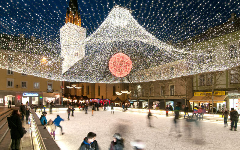 Schlittschuhlaufen auf einer dekorierten Eisbahn auf einem nächtlichen Stadtplatz, mit festlichen Lichtern und einem großen, beleuchteten roten Ornament über dem Kopf.
