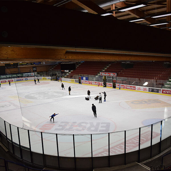 Mehrere Personen laufen Schlittschuh und spielen auf einer überdachten Eisbahn bei künstlicher Beleuchtung, die von leeren Tribünen umgeben ist.
