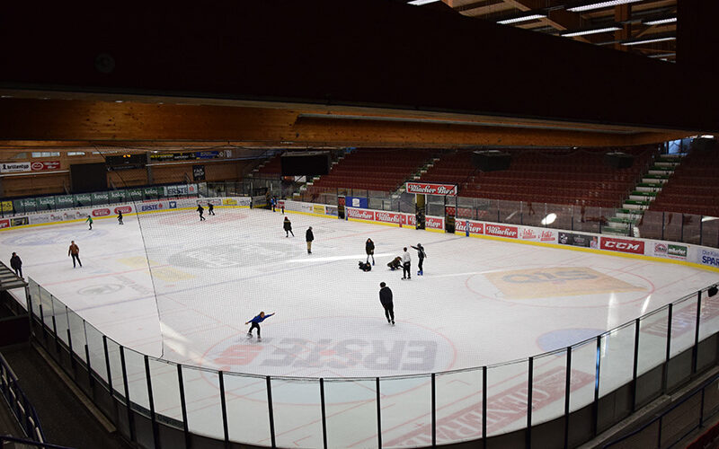 Mehrere Personen laufen Schlittschuh und spielen auf einer überdachten Eisbahn bei künstlicher Beleuchtung, die von leeren Tribünen umgeben ist.