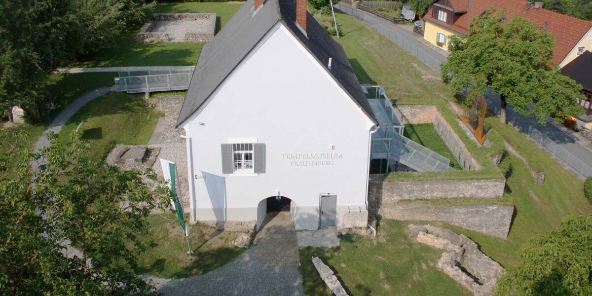 Luftaufnahme eines weißen Gebäudes mit der Aufschrift „Tempelmuseum Frauenberg“, umgeben von Steinruinen und Grünflächen, mit Häusern und einer Straße im Hintergrund.