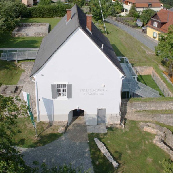Luftaufnahme eines weißen Gebäudes mit der Aufschrift „Tempelmuseum Frauenberg“, umgeben von Steinruinen und Grünflächen, mit Häusern und einer Straße im Hintergrund.