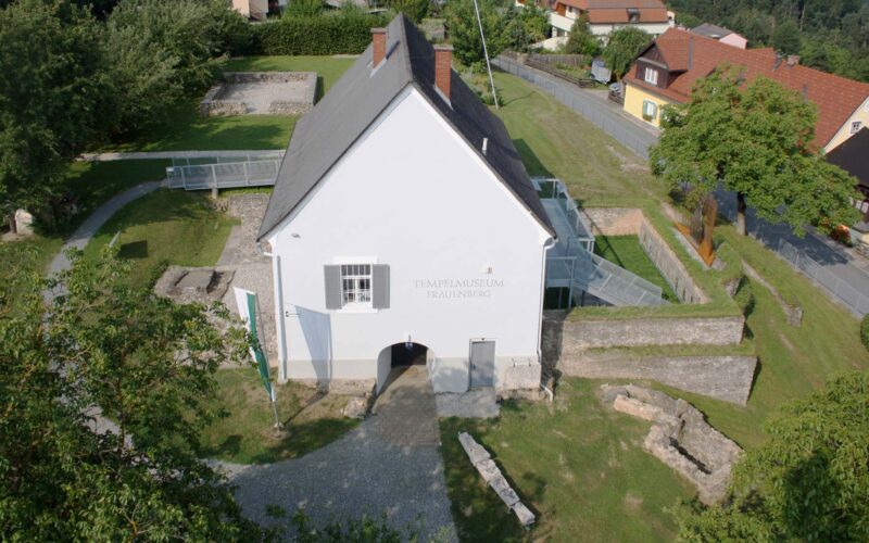 Luftaufnahme eines weißen Gebäudes mit der Aufschrift „Tempelmuseum Frauenberg“, umgeben von Steinruinen und Grünflächen, mit Häusern und einer Straße im Hintergrund.