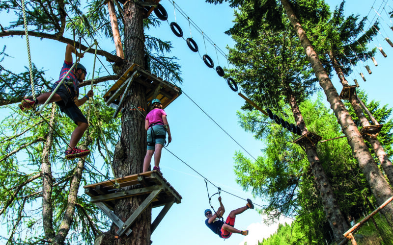 Drei Personen mit Helmen navigieren an einem sonnigen Tag durch einen Baumkronen-Seilgarten mit Plattformen, Seilen und aufgehängten Reifen zwischen hohen Bäumen.