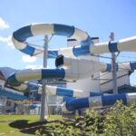 Eine große spiralförmige blaue und weiße Wasserrutsche im Freien steht neben einem Gebäude, mit Bergen und klarem Himmel im Hintergrund.