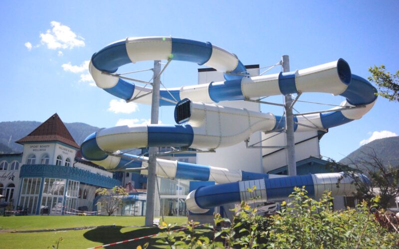 Eine große spiralförmige blaue und weiße Wasserrutsche im Freien steht neben einem Gebäude, mit Bergen und klarem Himmel im Hintergrund.