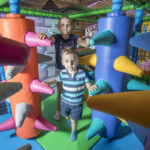 Zwei Kinder spielen auf einem Indoor-Spielplatz zwischen großen, gepolsterten Rollen mit bunten, weichen Stacheln in einem Spielbereich mit Netzen.