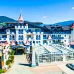 Ein großes, schlossähnliches Hotel mit Türmen und roten Dächern, umgeben von Bergen und grünen Feldern unter einem strahlend blauen Himmel, mit geparkten Autos davor.
