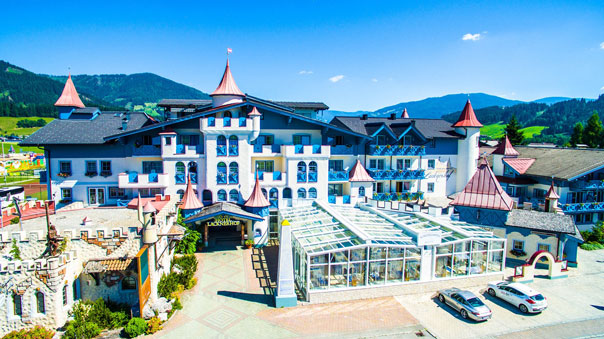 Ein großes, schlossähnliches Hotel mit Türmen und roten Dächern, umgeben von Bergen und grünen Feldern unter einem strahlend blauen Himmel, mit geparkten Autos davor.