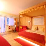 Ein Hotelzimmer mit Holzwänden, einem Himmelbett, rotem Bettzeug, einem Schreibtisch, einem Sofa, einem kleinen Tisch mit Stühlen und einer Balkontür, durch die Tageslicht einfällt.