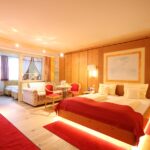 Ein geräumiges Hotelzimmer mit einem Doppelbett, roten Akzenten, Holzwänden, Sitzecke und einem angrenzenden kleineren Zimmer mit zwei Einzelbetten.