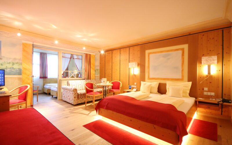 Ein geräumiges Hotelzimmer mit einem Doppelbett, roten Akzenten, Holzwänden, Sitzecke und einem angrenzenden kleineren Zimmer mit zwei Einzelbetten.