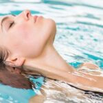 Eine Frau in einem schwarzen Badeanzug schwimmt auf dem Rücken in einem Schwimmbecken, das Gesicht nach oben geneigt und die Augen geschlossen.