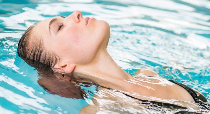 Eine Frau in einem schwarzen Badeanzug schwimmt auf dem Rücken in einem Schwimmbecken, das Gesicht nach oben geneigt und die Augen geschlossen.