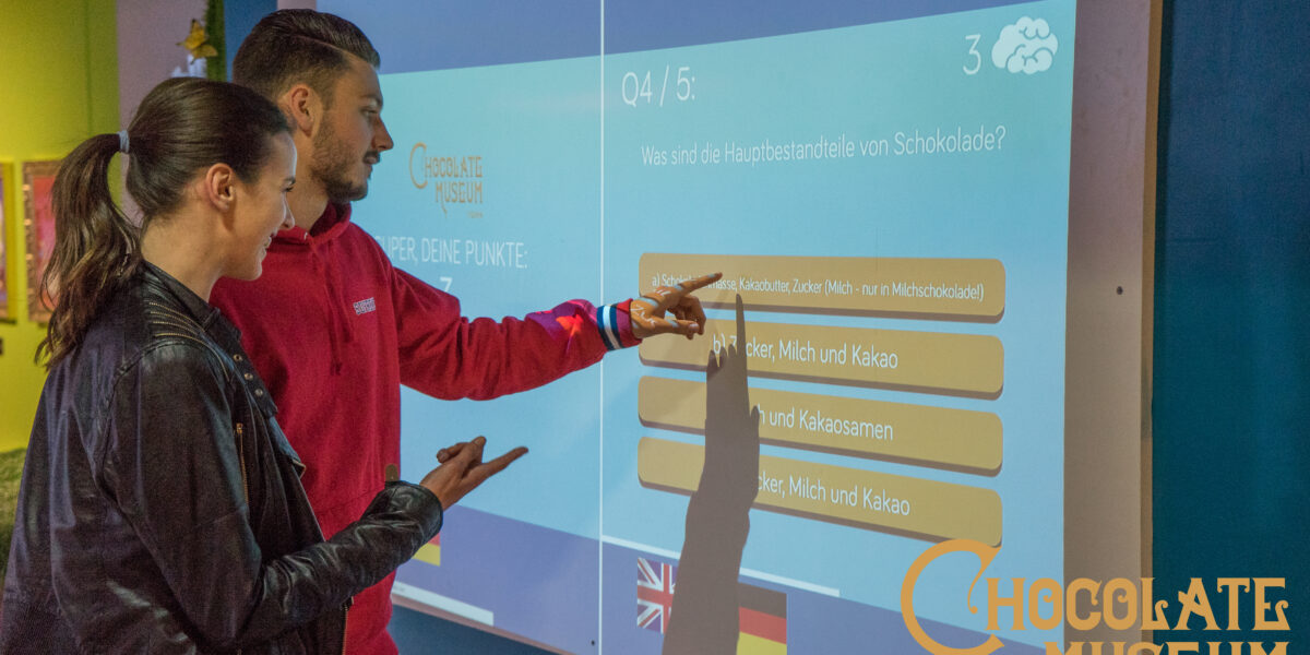 Zwei Personen interagieren mit einem Touchscreen-Quiz über Schokolade im Schokoladenmuseum Wien, wobei die Antwortmöglichkeiten auf Deutsch und Englisch sichtbar sind.