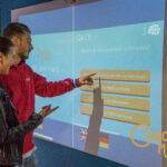 Zwei Personen interagieren mit einem Touchscreen-Quiz über Schokolade im Schokoladenmuseum Wien, wobei die Antwortmöglichkeiten auf Deutsch und Englisch sichtbar sind.