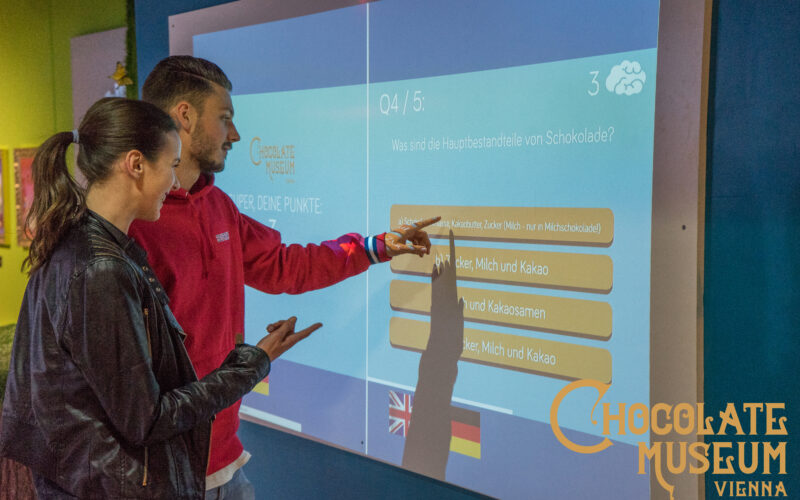 Zwei Personen interagieren mit einem Touchscreen-Quiz über Schokolade im Schokoladenmuseum Wien, wobei die Antwortmöglichkeiten auf Deutsch und Englisch sichtbar sind.