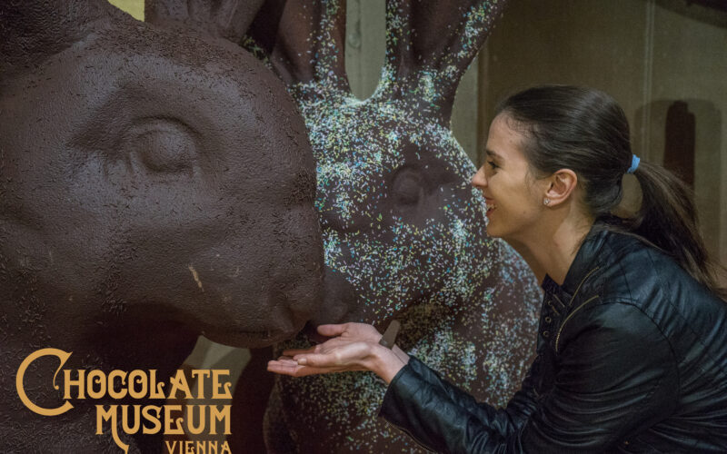 Eine Frau lächelt und posiert mit einer großen Schokoladenhasen-Statue im Schokoladenmuseum Wien.