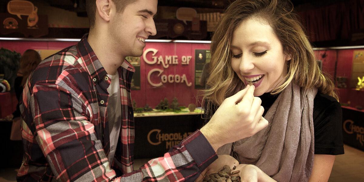 Ein Mann hält Kakaobohnen in der Hand, während eine Frau lächelnd eine isst; sie stehen vor einem Schild mit der Aufschrift "Game of Cocoa".