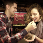 Ein Mann hält Kakaobohnen in der Hand, während eine Frau lächelnd eine isst; sie stehen vor einem Schild mit der Aufschrift "Game of Cocoa".