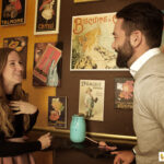 Eine Frau und ein Mann lächeln und unterhalten sich vor einer mit alten Schokoladen- und Keksplakaten dekorierten Wand. Eine blaue Dose steht auf der Theke zwischen ihnen.