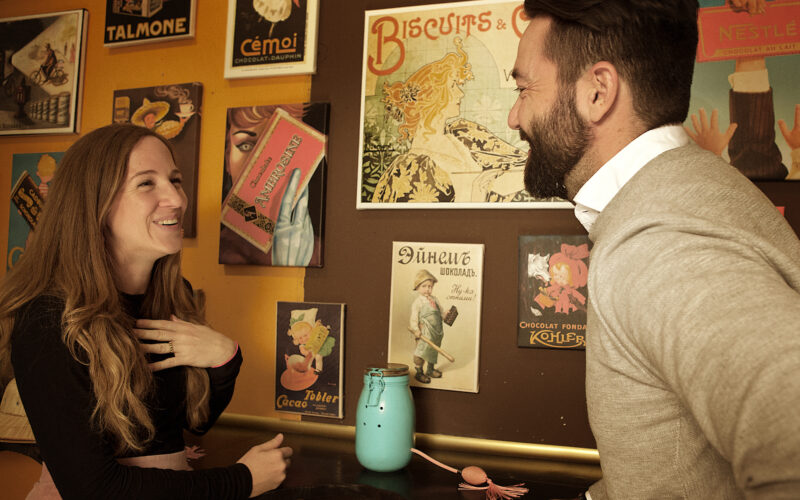 Eine Frau und ein Mann lächeln und unterhalten sich vor einer mit alten Schokoladen- und Keksplakaten dekorierten Wand. Eine blaue Dose steht auf der Theke zwischen ihnen.