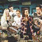 Eine Gruppe von sechs Personen versammelt sich um eine Theke mit verschiedenen Pralinen, probiert und diskutiert über die Leckereien in einer Ladenumgebung.