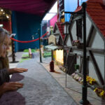 Zwei Mädchen betrachten in einem Museum oder Ausstellungsraum ein detailliertes Miniaturmodell einer Straße mit Gebäuden, Blumen und einem hellen Schaufenster.