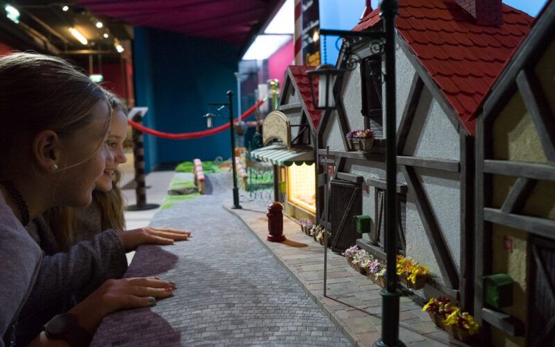 Zwei Mädchen betrachten in einem Museum oder Ausstellungsraum ein detailliertes Miniaturmodell einer Straße mit Gebäuden, Blumen und einem hellen Schaufenster.