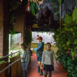 Drei Kinder betrachten Ausstellungsstücke im Schokoladenmuseum Wien mit viel Grün und einem großen Gorillabild im Hintergrund.